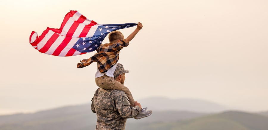 A man in military uniform with little boy on his shoulders waving an American flag